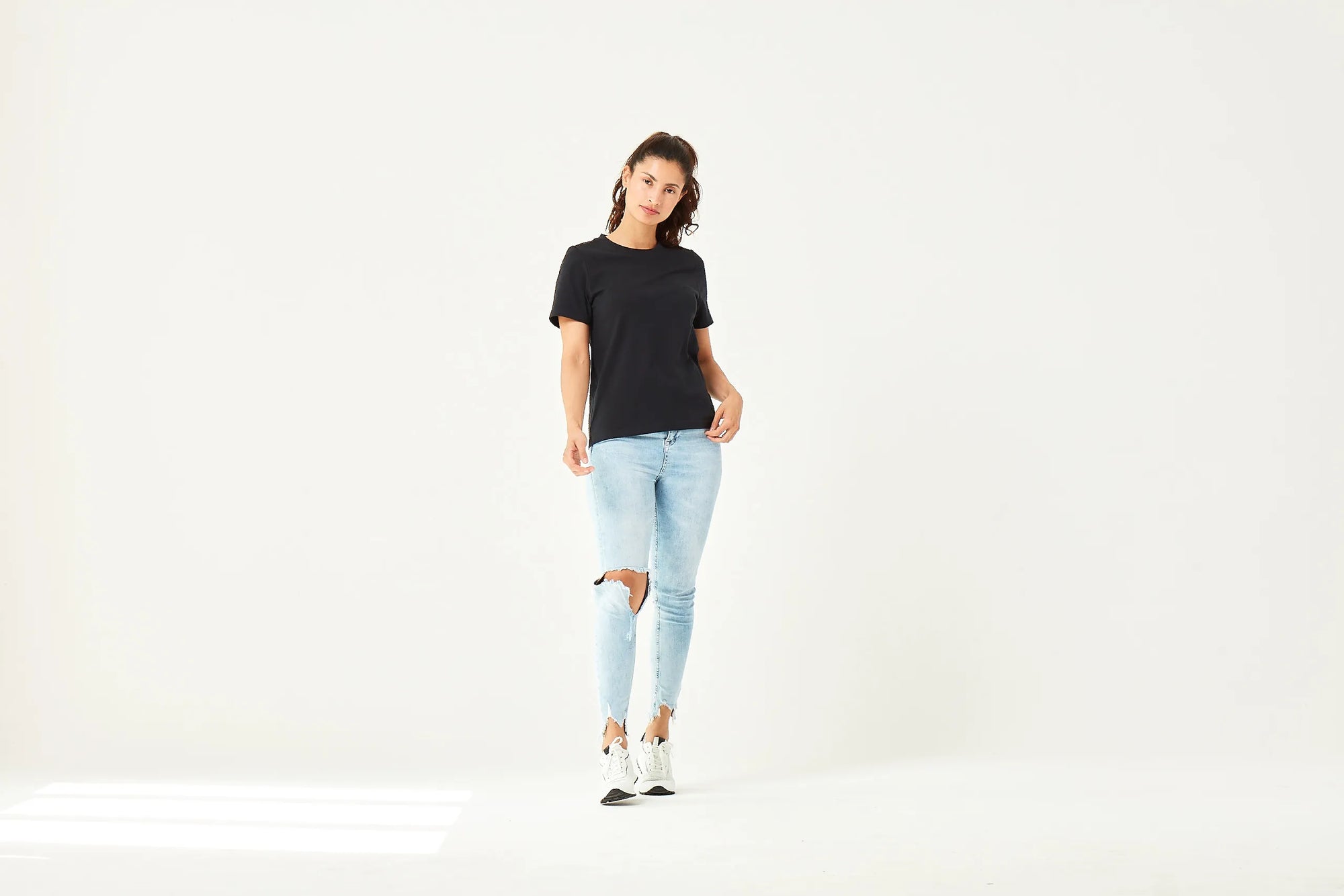 Frau in schwarzem kurzärmligen T-Shirt, hellblauen zerrissenen Jeans und weißen Sneakers mit schwarzen Akzenten, stehend vor weißem Hintergrund.