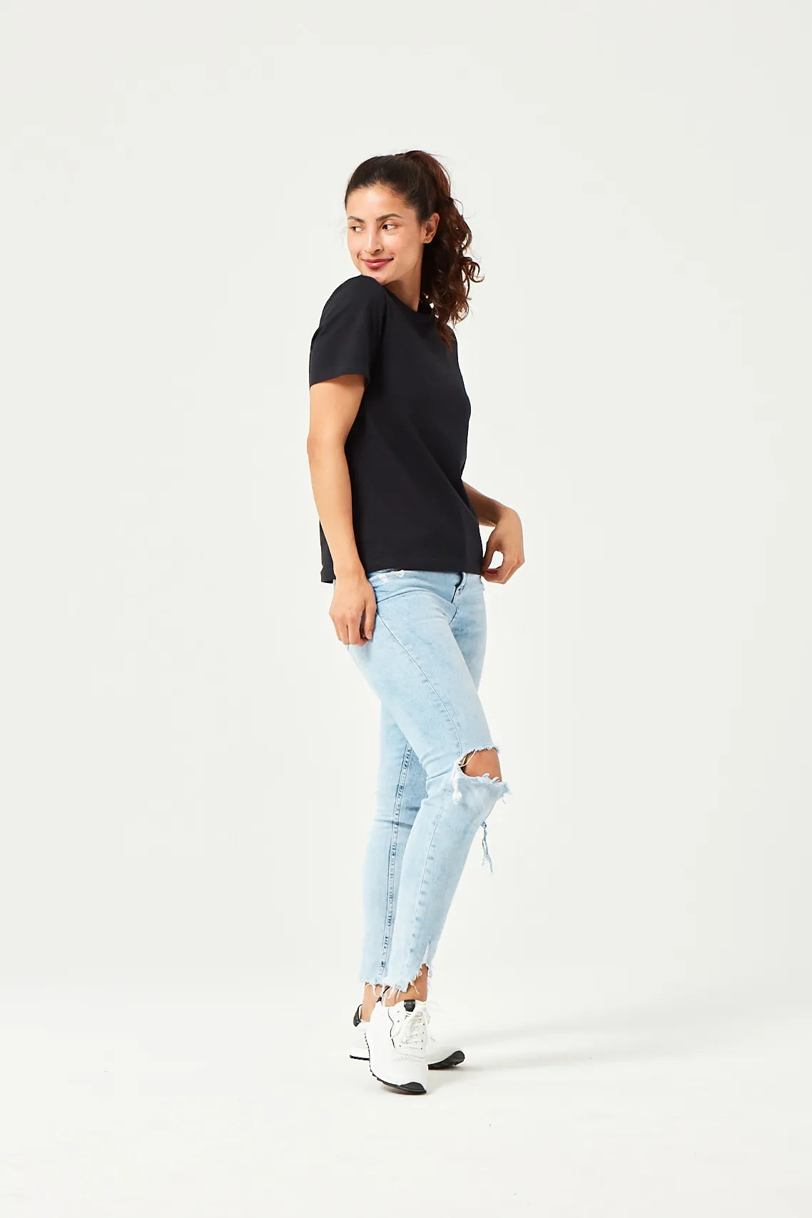 Frau in schwarzem kurzärmligen T-Shirt, hellblauen zerrissenen Jeans und weißen Sneakers mit schwarzen Akzenten, stehend vor weißem Hintergrund.