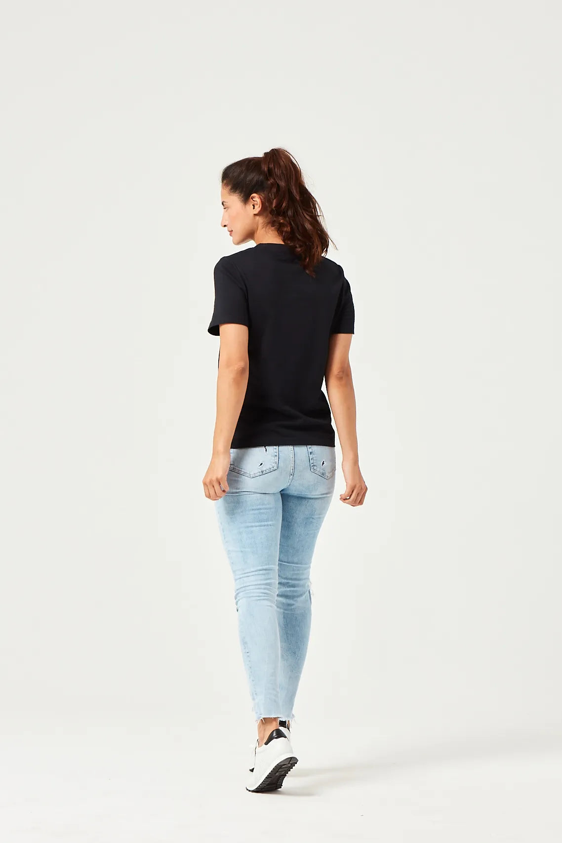 Frau in schwarzem kurzärmligen T-Shirt, hellblauen zerrissenen Jeans und weißen Sneakers mit schwarzen Akzenten, stehend vor weißem Hintergrund.