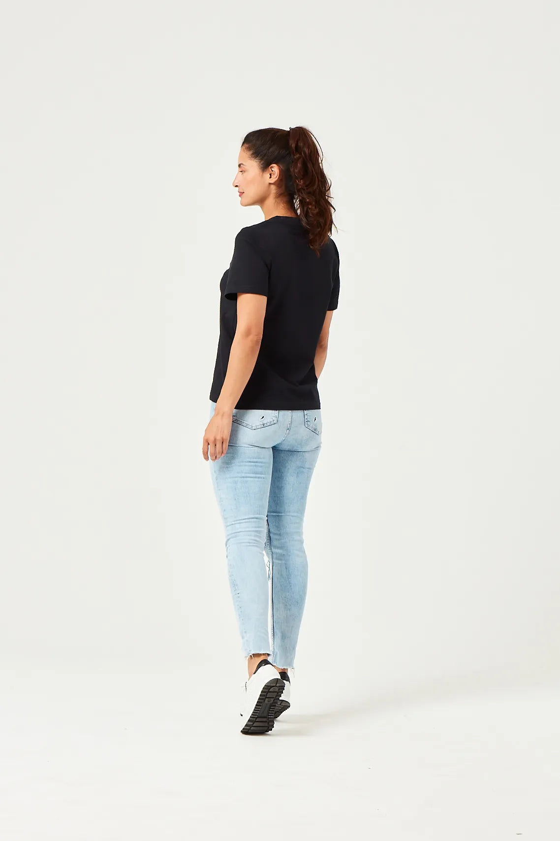 Rückansicht einer Frau in einem schwarzen T-Shirt, hellblauen Jeans und weißen Sneakers auf weißem Hintergrund, Haar zu einem Pferdeschwanz gebunden.